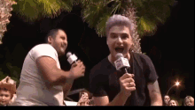 a man with purple hair is singing into a microphone while another man stands behind him holding a microphone