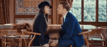a man in a suit and a woman in a hat are sitting at a table holding hands .
