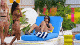 a woman in a bathing suit sits on a blue lounge chair while another woman walks behind her