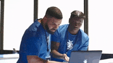 two men in blue shirts are looking at a laptop together .