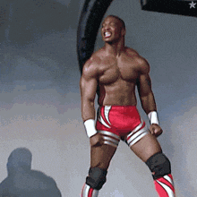 a shirtless wrestler wearing red and white shorts and knee pads stands with his fist in the air