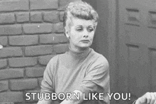 a black and white photo of a woman sitting in front of a brick wall saying `` stubborn like you '' .