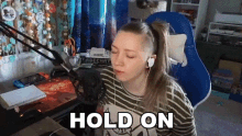 a woman wearing headphones and a striped shirt is sitting in front of a microphone and says `` hold on '' .