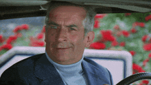 a man in a suit and turtleneck is sitting in a car with red flowers in the background