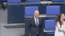a man in a suit and tie stands in front of a row of blue chairs with 2df on the bottom right