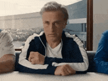 a man in a blue and white track jacket is sitting at a table with his hands folded .