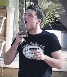 a man wearing a t-shirt that says clermont lounge is holding a cell phone to his mouth .