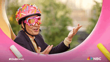 a man wearing a helmet with sprinkles on it is standing in front of a pink donut with the nbc logo