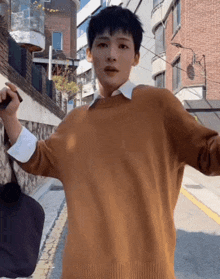 a young man wearing a brown sweater is standing on a street