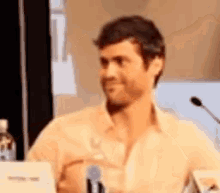 a man in a tan shirt is sitting at a table with a microphone .