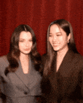 two women standing next to each other with a red curtain behind them