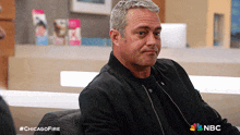 a man in a black jacket is sitting on a couch with a nbc logo behind him