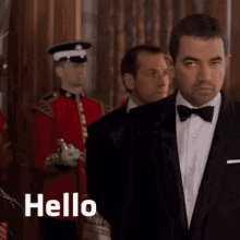 a man in a tuxedo and bow tie says hello in front of a group of men in uniforms