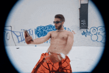 a shirtless man in orange shorts stands in front of a wall with graffiti on it including one that says hi