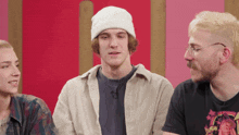 a man wearing a white beanie and glasses talks to two other people