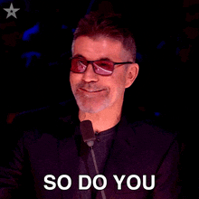 a man wearing glasses and a suit says so do you in front of a microphone
