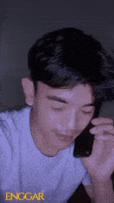 a young man in a white shirt is talking on a cell phone with the word enggar on the bottom