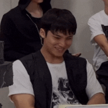 a man wearing a black vest and a white shirt with the letter l on it smiles