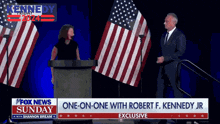 a woman stands at a podium talking to a man in front of american flags with a fox news sunday banner behind her