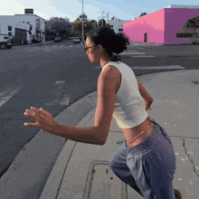 a woman in a white tank top and blue pants stands on a sidewalk near a pink building