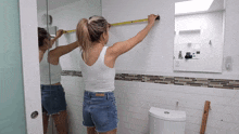 a woman is measuring a bathroom wall with a tape measure