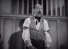 a man in a vest and bow tie is standing in front of a wooden wall with his mouth open