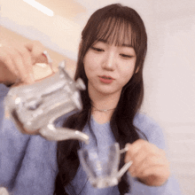 a girl in a blue sweater is pouring something into a glass from a teapot
