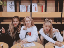 three girls are sitting at a table in front of a bookshelf and making funny faces .