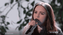 a woman singing into a microphone with a kat hammock logo in the background