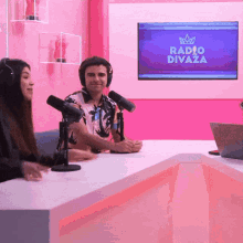 a man and a woman are sitting at a table with microphones in front of a screen that says radio divaza