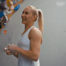 a woman in a white tank top is smiling in front of a wall with the olympic rings on it