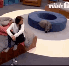 a woman is sitting on a bed in front of a blue ottoman that says milele