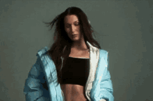 a woman wearing a blue jacket and a black crop top is standing in front of a grey background .