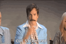 a man with a mustache is sitting in front of a group of people with the words cue stephanie .