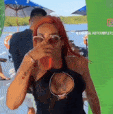 a woman with red hair and sunglasses is drinking a drink while covering her nose .