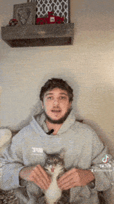a man in a grey hoodie is holding a cat while talking into a microphone