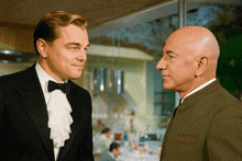 a man in a tuxedo stands next to a bald man in a green jacket