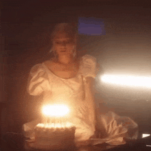 a woman in a white dress is sitting at a table with a birthday cake with candles on it .