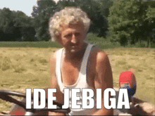 a man in a white tank top is sitting in a field with the words ide jebiga on the bottom