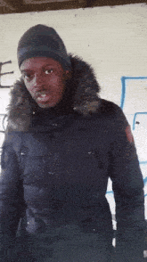 a man wearing a black jacket with a fur hood is standing in front of a wall with the letter e painted on it