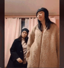 two women are standing next to each other in a room . one of the women is wearing a beanie .