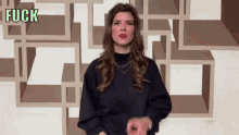a woman in a black sweater says fuck in front of a patterned wall