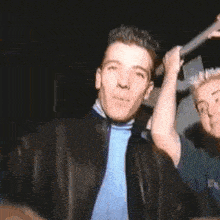 a man in a black jacket and blue tie is standing next to another man holding a microphone