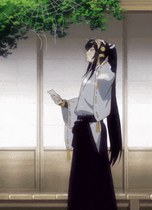 a man with long black hair is standing in front of a window holding a piece of paper