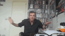 a man is sitting at a desk with his arms outstretched in front of a wall of dvds .