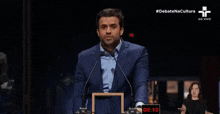 a man in a suit and tie is giving a speech in front of two microphones while a woman looks on