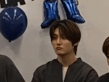 a young man is sitting in front of a group of people with balloons on the wall behind him .