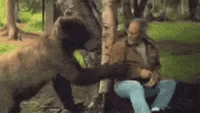 a man is sitting next to a tree in the woods while a bear attacks him .