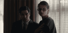 a man in a suit and tie stands next to a woman wearing glasses and earrings with a netflix logo in the corner