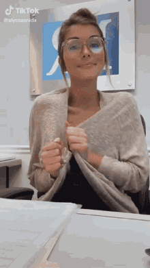 a woman wearing glasses and a sweater is sitting at a desk in front of a sign that says " tiktok "